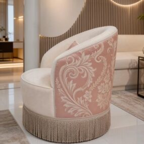 A cream barrel accent chair with a pink damask-patterned back panel and long bullion fringe skirt on a glossy tile floor.