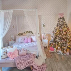 A pink girls’ holiday bedroom with a white canopy bed, pink pillows and chunky knit throw, and a flocked Christmas tree decorated in pink ornaments.