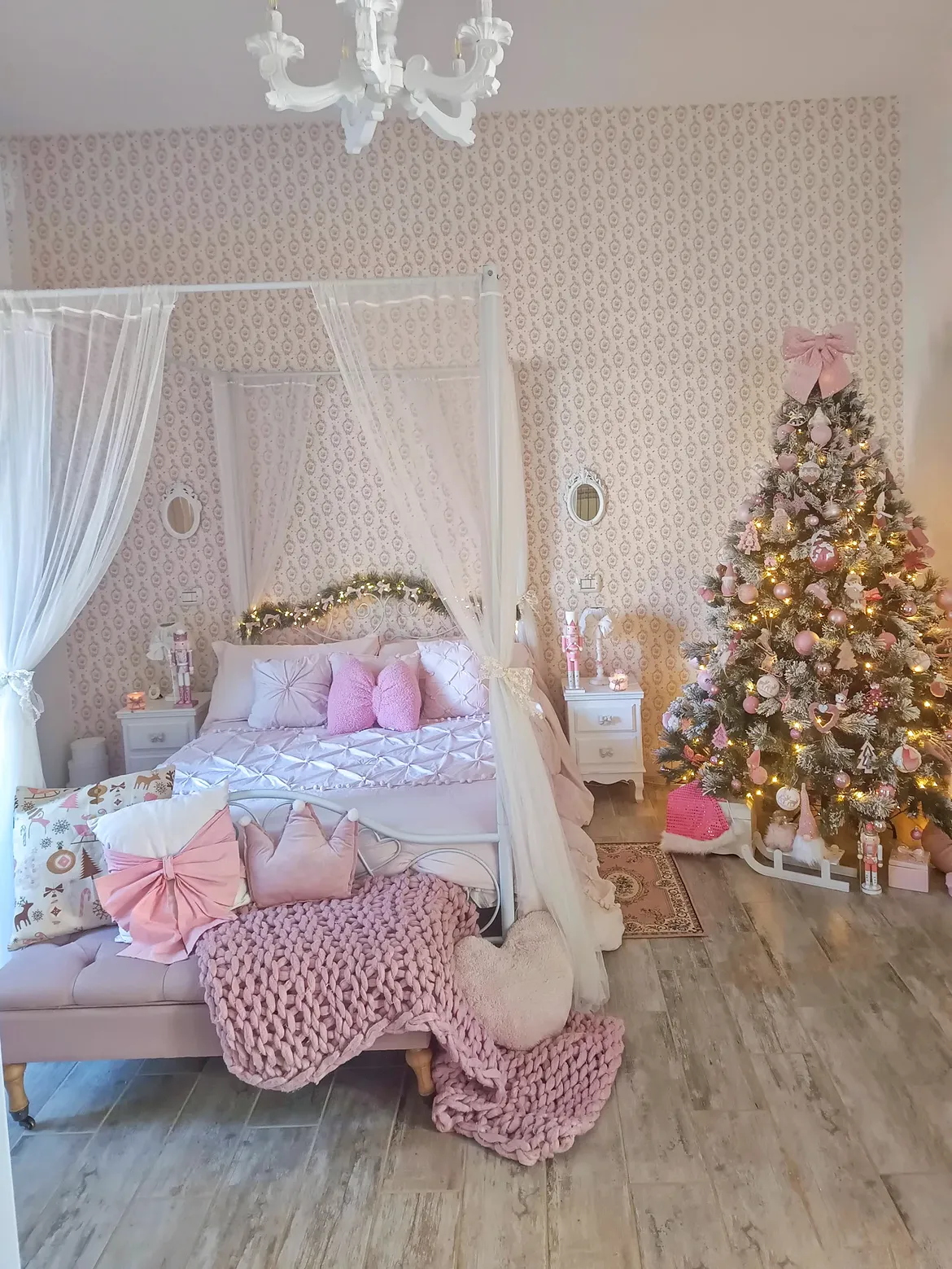 A pink girls’ holiday bedroom with a white canopy bed, pink pillows and chunky knit throw, and a flocked Christmas tree decorated in pink ornaments.