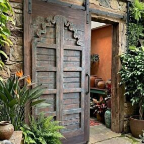 A carved wood sliding barn door with iron hardware set into a stone wall, surrounded by ferns, bird of paradise, and clay pots.