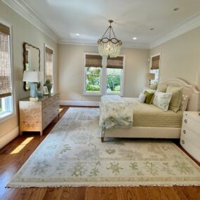 A cream upholstered bed with sage green bedding, a burl wood dresser, soft green floral rug, and a beaded chandelier in a bright bedroom.