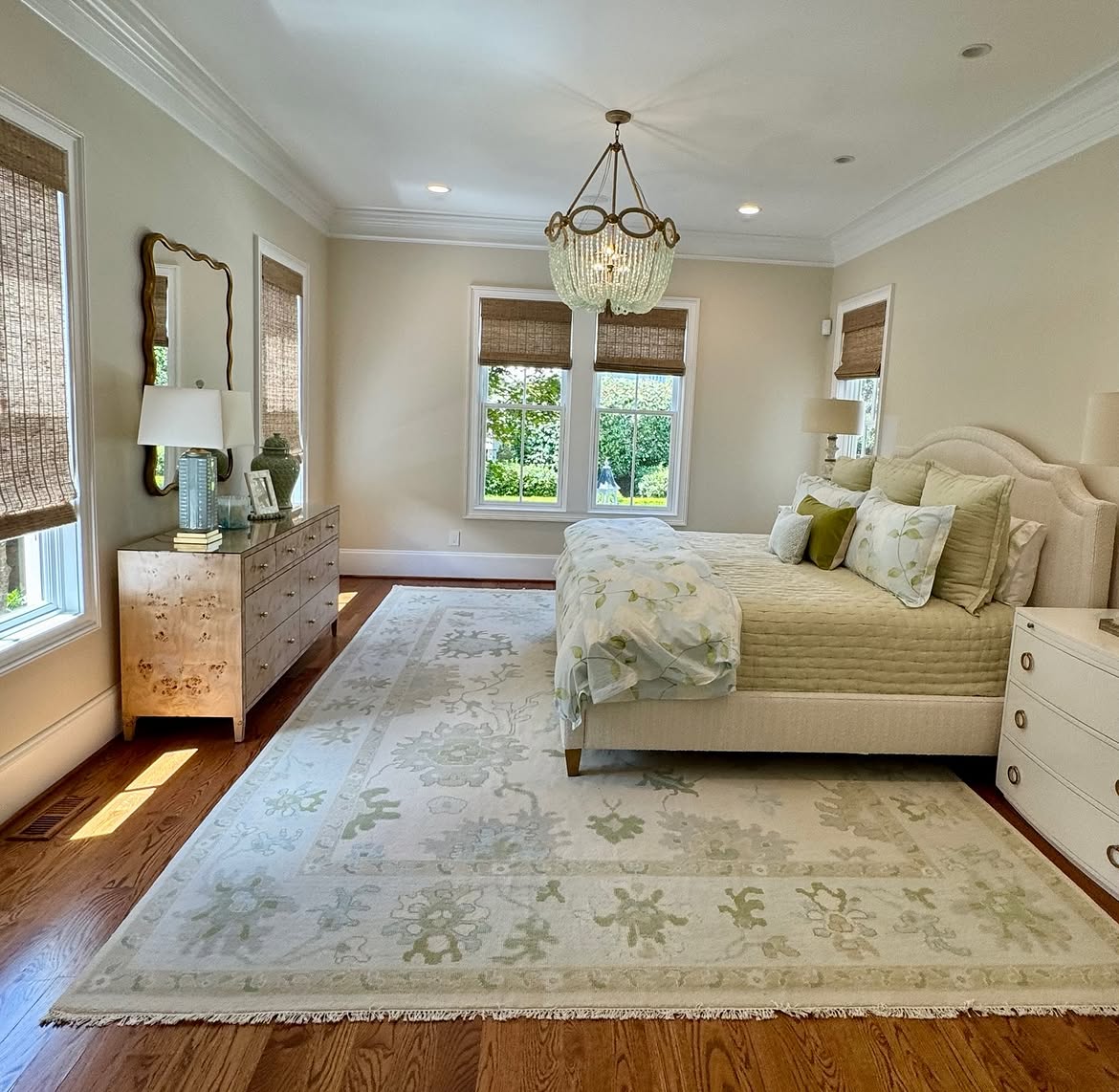 A cream upholstered bed with sage green bedding, a burl wood dresser, soft green floral rug, and a beaded chandelier in a bright bedroom.