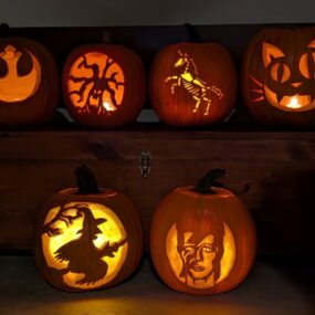 Six carved and lit jack-o-lanterns displayed on and in front of a wooden chest, featuring a witch flying past the moon, a cheshire cat, a skeleton horse, and other detailed designs.
