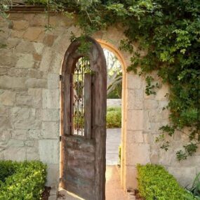 An open arched weathered wood garden gate with an iron grille window set into a stone wall draped with ivy and flanked by boxwoods.