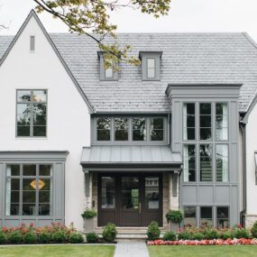 A modern Tudor-style home with white stucco, gray board and batten siding, slate shingle roof, dark wood double front door, bay windows, and manicured landscaping with hydrangeas.