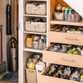 Under-stair storage with pull-out shoe drawers, a tall cleaning closet with hung tools, and open shelves of baskets and bottles.