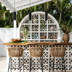 An outdoor tropical bar with a white breeze-block arched back wall, live-edge wood counter, and three rope-woven stools under a pergola.