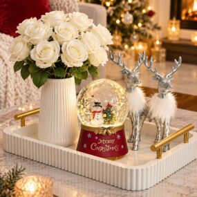 A fluted white tray on a marble coffee table styled with white roses, a Merry Christmas snow globe, and two silver feathered reindeer figurines.
