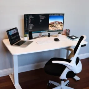 A minimalist white standing desk with an ultrawide curved monitor, open laptop, white keyboard, black and white mesh office chair, and small desk accessories on a dark wood floor.